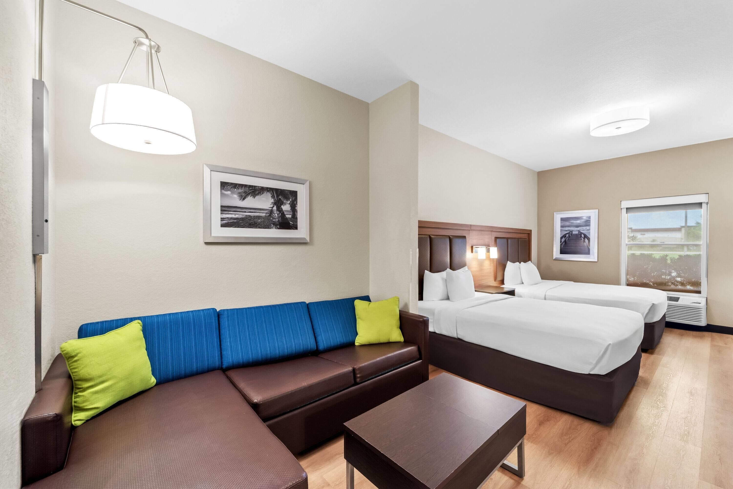 Quarto de hotel com sofá marrom, cama com roupa de cama branca, uma mesinha e um quadro na parede.