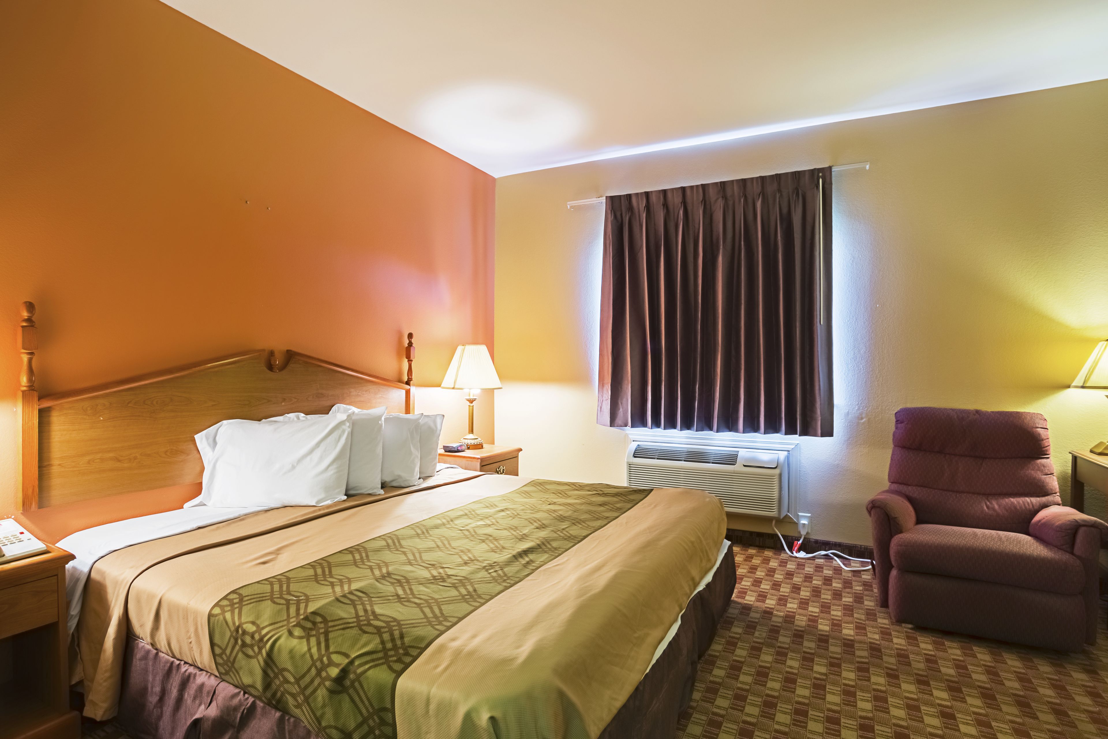 A hotel room with a bed, a chair, a nightstand, a lamp, and an air conditioning unit.