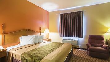 A hotel room with a bed, a chair, a nightstand, a lamp, and an air conditioning unit.