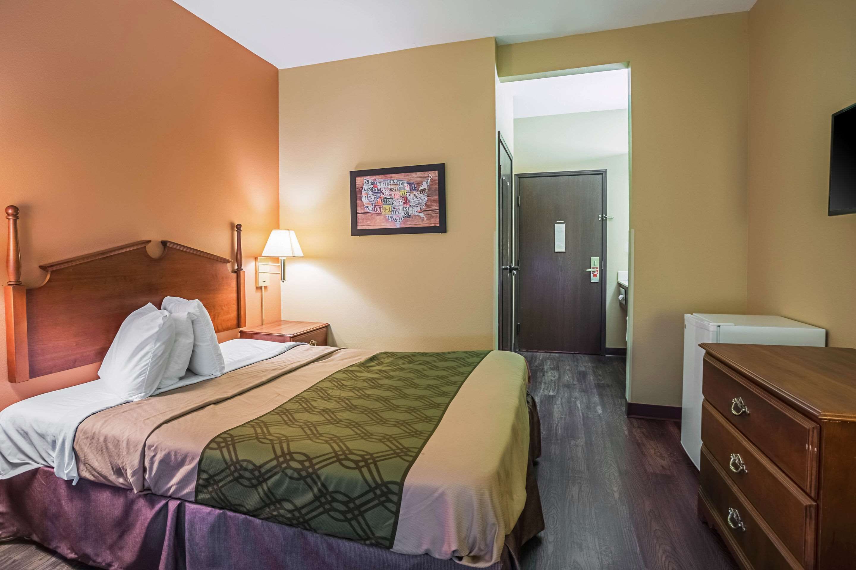 A hotel room with a wooden bed, a nightstand with a lamp, a dresser, and a refrigerator.