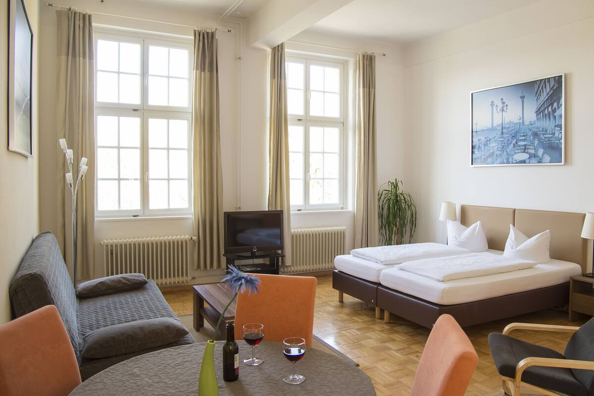 A hotel room with a bed, a desk, a chair, a table with a bottle and glass of wine, and a painting on the wall.