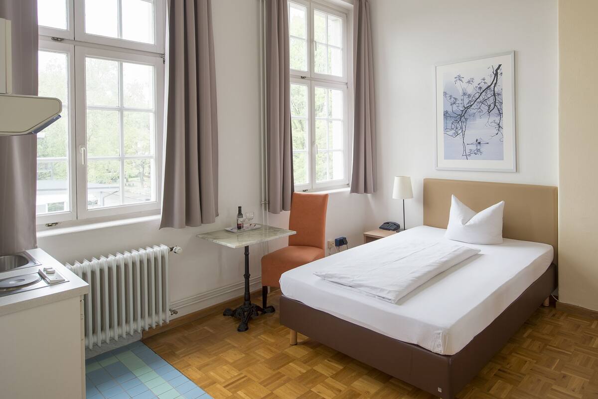 A hotel room with a bed, a table with a lamp, a chair, a radiator, and a window with curtains.