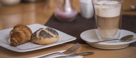 A table with a croissant, a poppy seed roll, and a glass of coffee with foam.