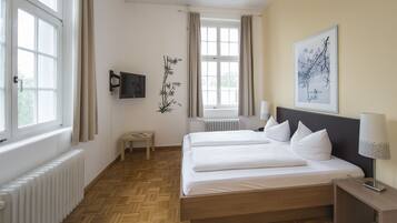 A hotel room with two beds, a TV, a small table, a radiator, and a window with curtains.