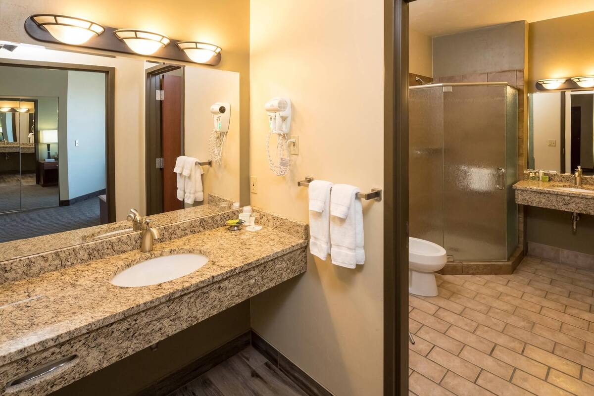 A hotel bathroom with a granite countertop sink, a large mirror, two hair dryers, a toilet, and a glass-enclosed shower.
