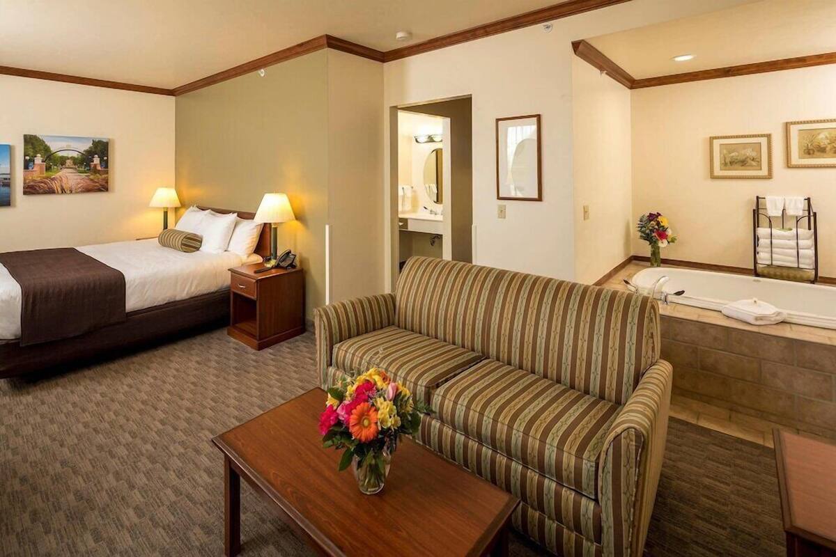 A hotel room with a bed, a sofa, a small table with a vase of flowers, a bathroom with a bathtub, and a mirror.