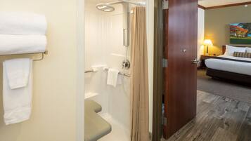 A hotel room with a shower, a toilet, a bed, and a nightstand with a lamp.