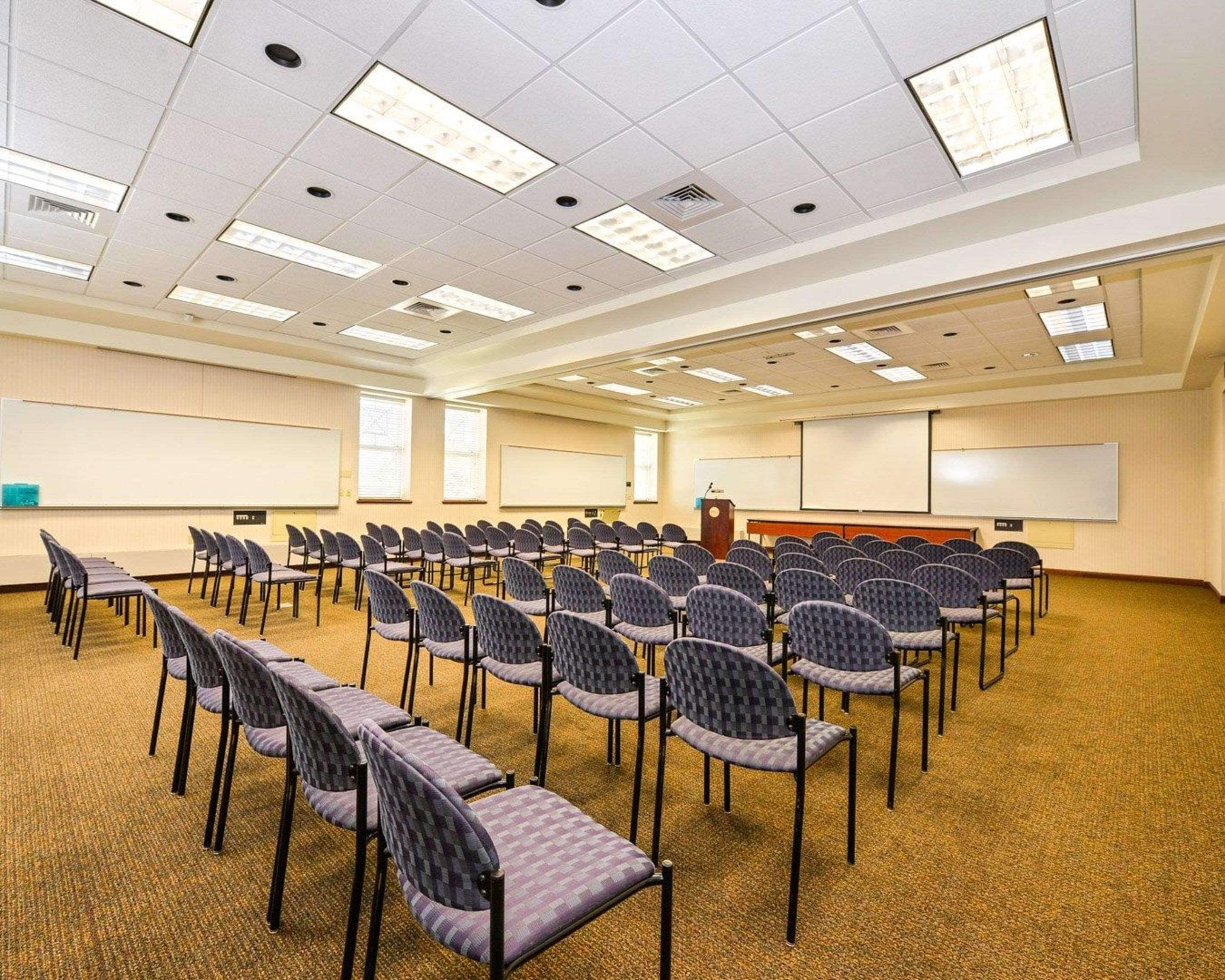 A conference room with rows of chairs, a podium, a projector screen, and a carpeted floor.