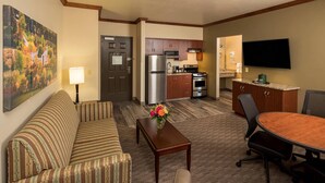 A hotel room with a sofa, a dining table with chairs, a kitchen area with a refrigerator, stove, microwave, and a flat-screen TV mounted on the wall. - Kress Inn, an Ascend Collection Hotel (De Pere)