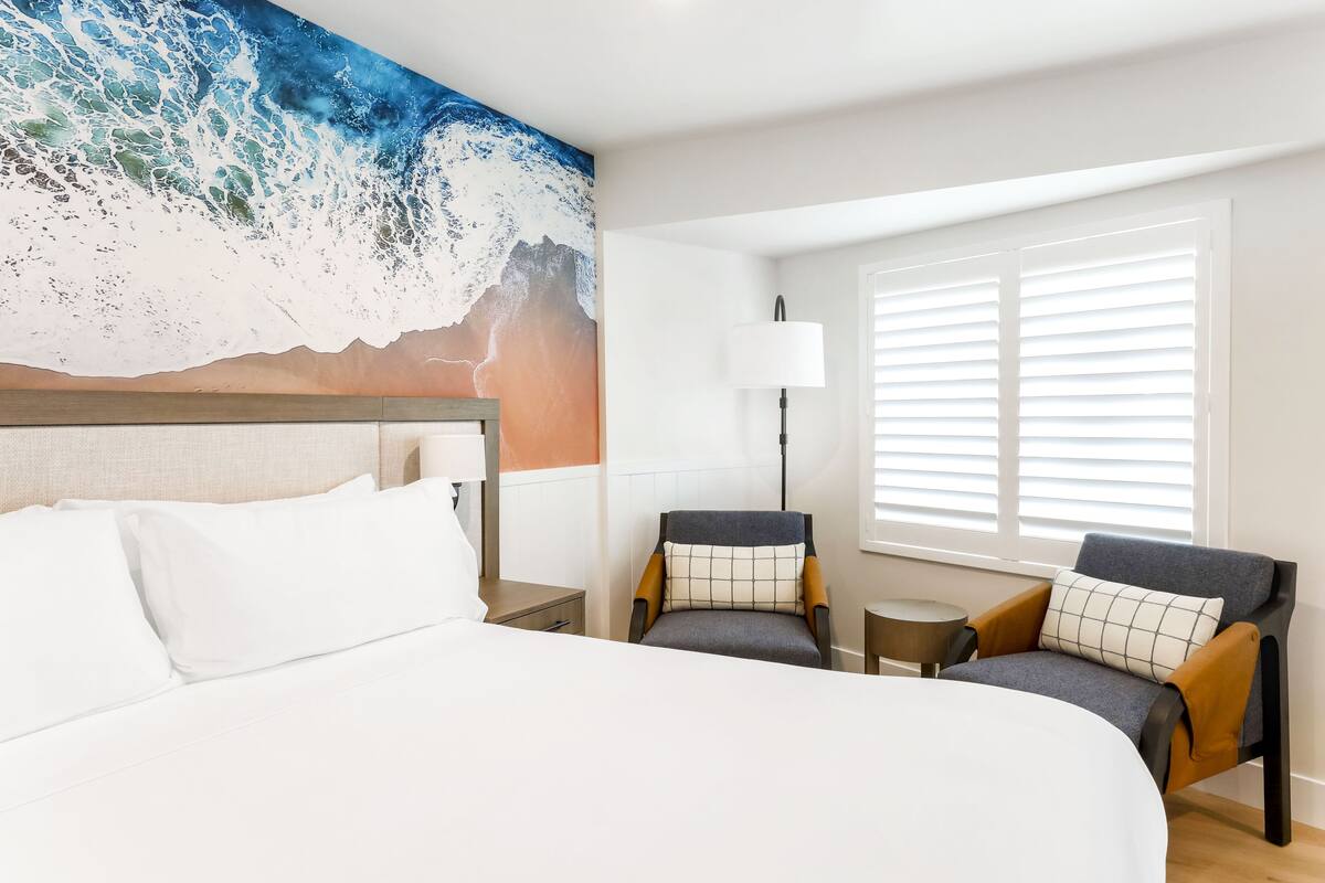 A hotel room with a large bed, two armchairs, a small table, a lamp, and a window with blinds.