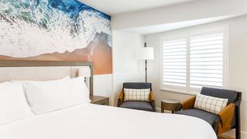 A hotel room with a large bed, two armchairs, a small table, a lamp, and a window with blinds.