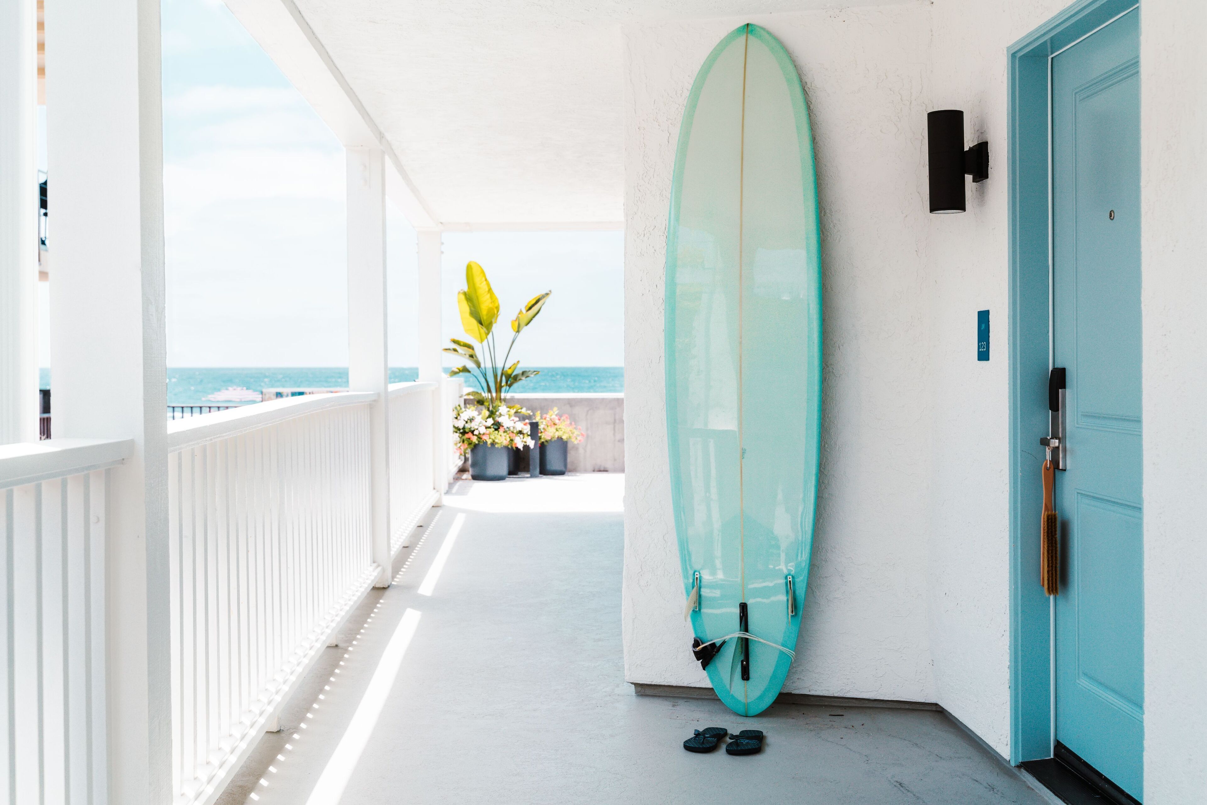 A balcony with a surfboard leaning against the wall, a door, and potted plants.