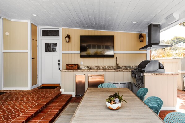 A modern kitchen with a flat-screen TV, built-in appliances, and a dining area with a wooden table and blue chairs. - Del Mar Beach Hotel (Del Mar)