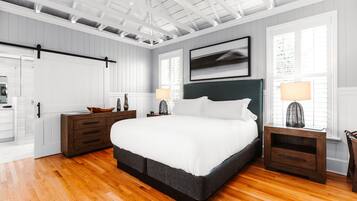 Une chambre avec un grand lit, un plancher en bois, des murs blancs et un plafond avec des poutres apparentes.