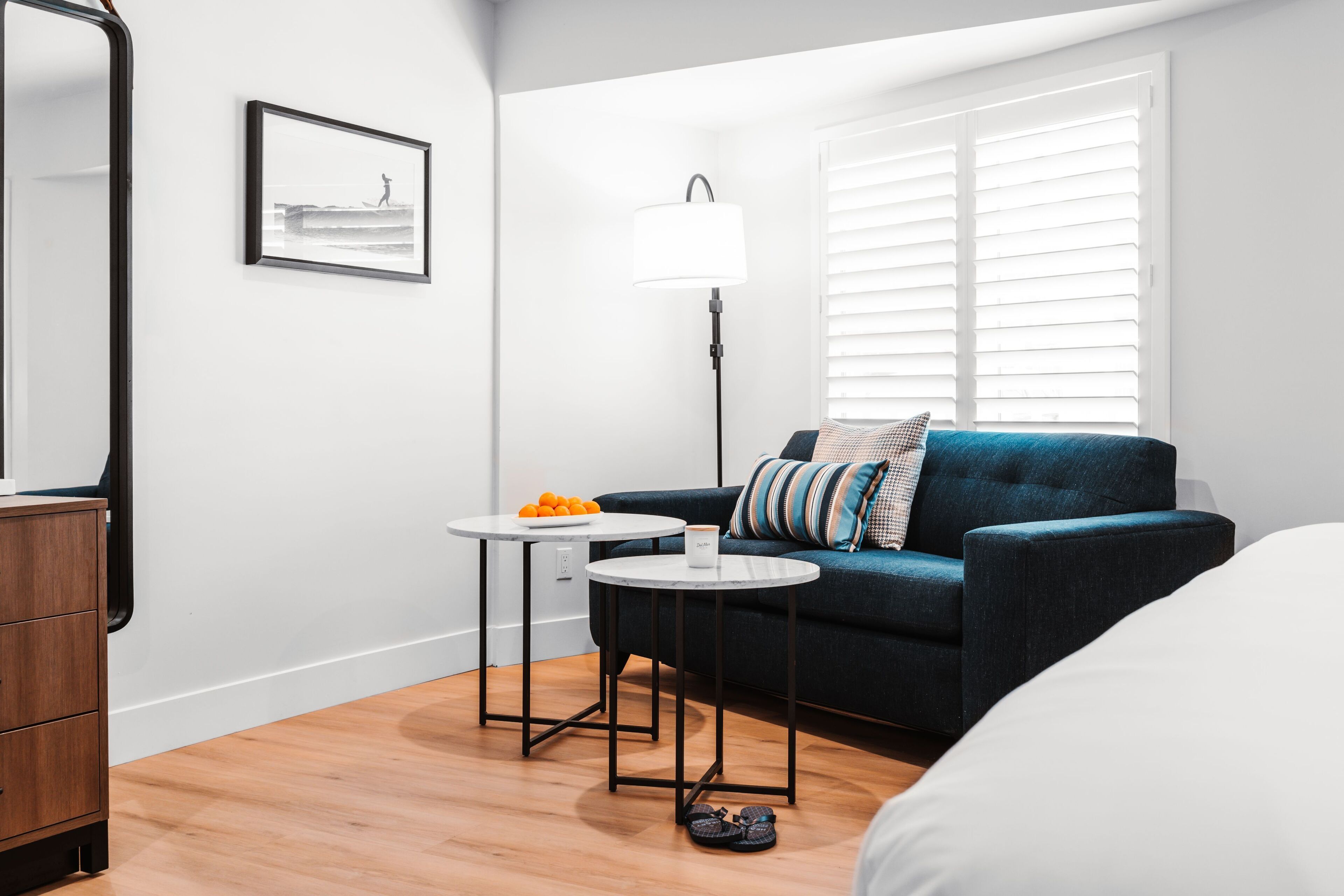 A modern hotel room with a blue sofa, a small round table with a lamp, and a framed picture on the wall.