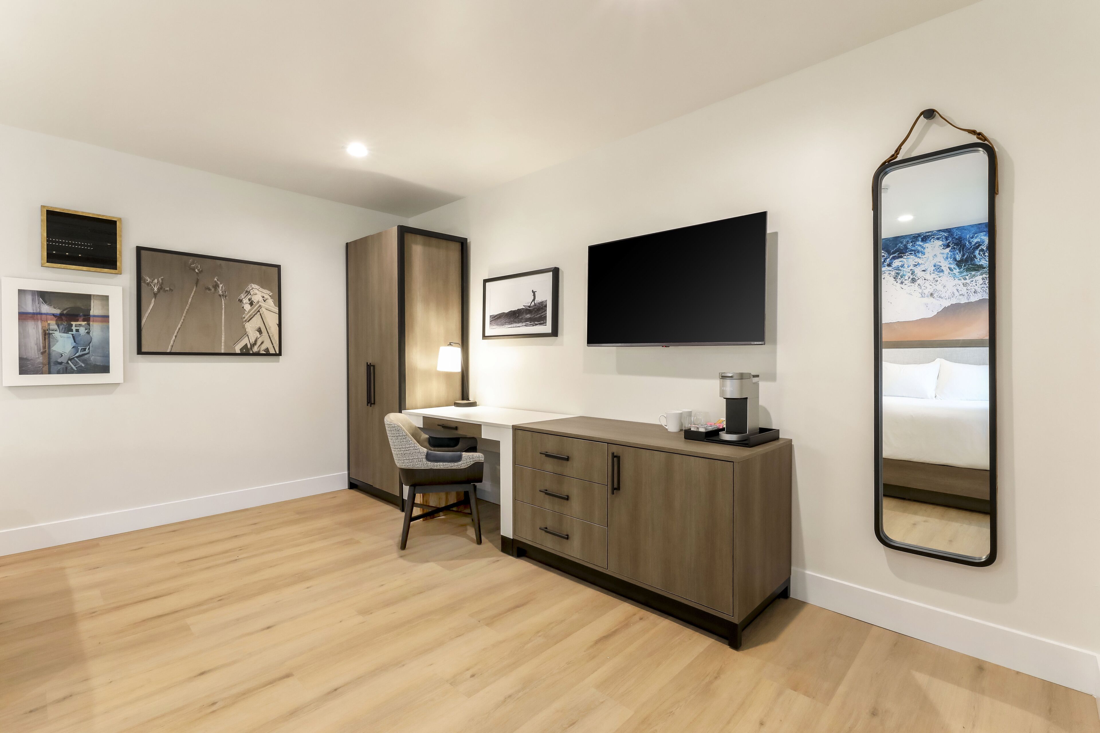 A modern bedroom with a wooden floor, a desk with a chair, a bed, a TV, and artwork on the walls.