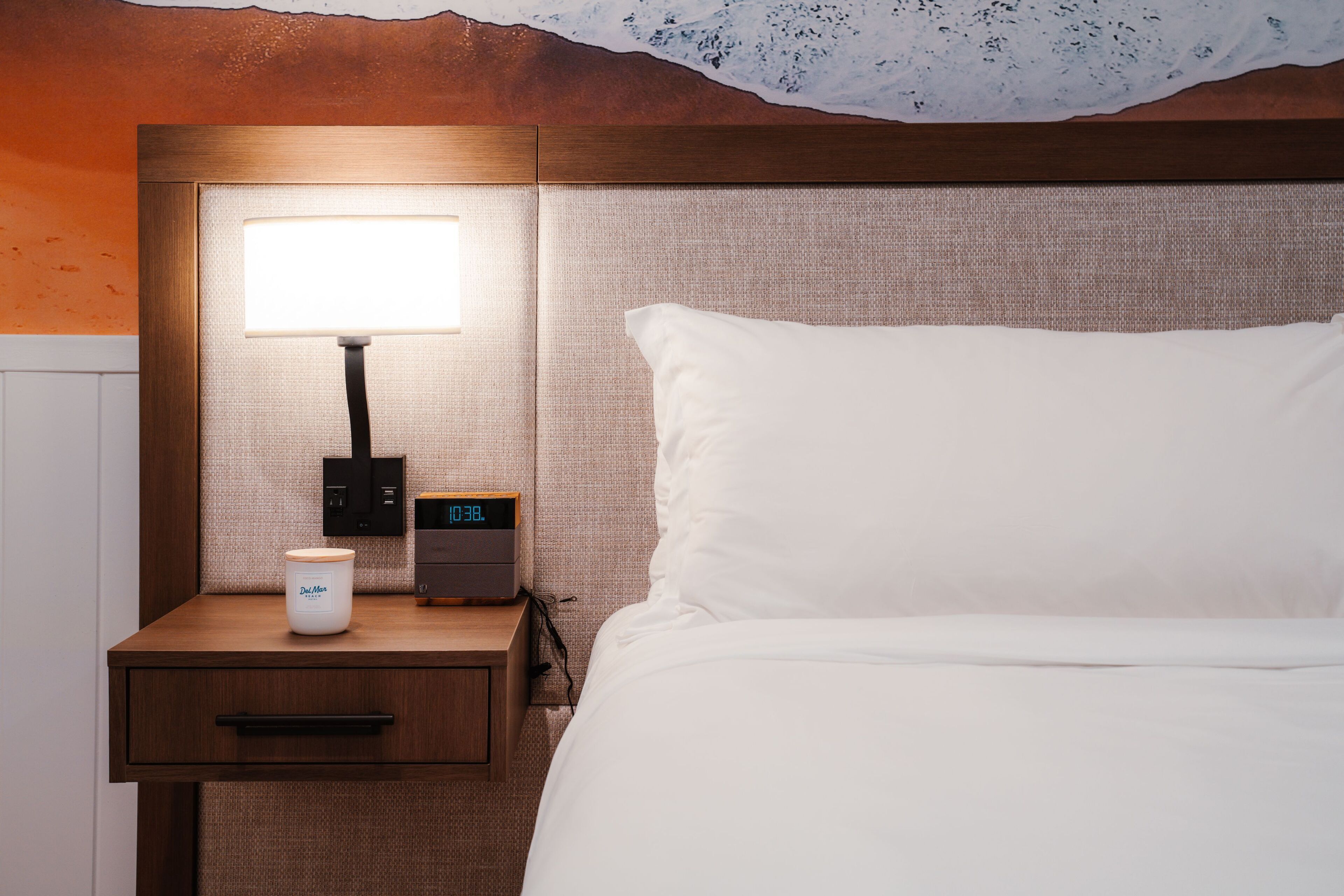 A neatly made bed with white linens, a bedside table with a lamp, a digital clock, and a small container.