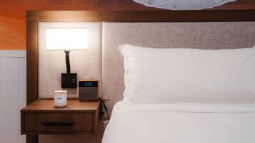 A neatly made bed with white linens, a bedside table with a lamp, a digital clock, and a small container.