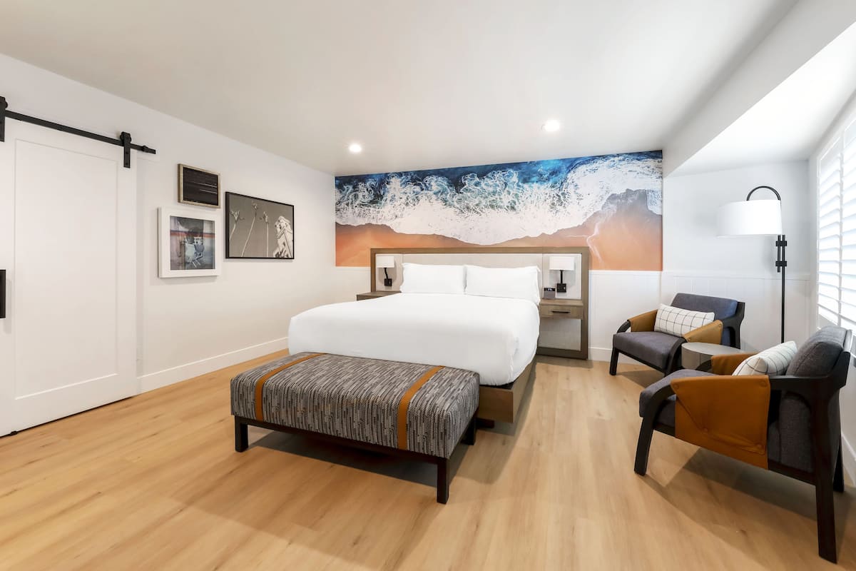 A modern bedroom with a large bed, a bench, two armchairs, a nightstand, and a wall-mounted lamp.