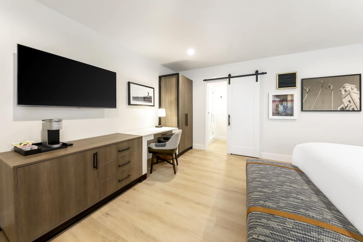 A modern hotel room with a large bed, a desk with a coffee maker, a chair, and artwork on the walls.