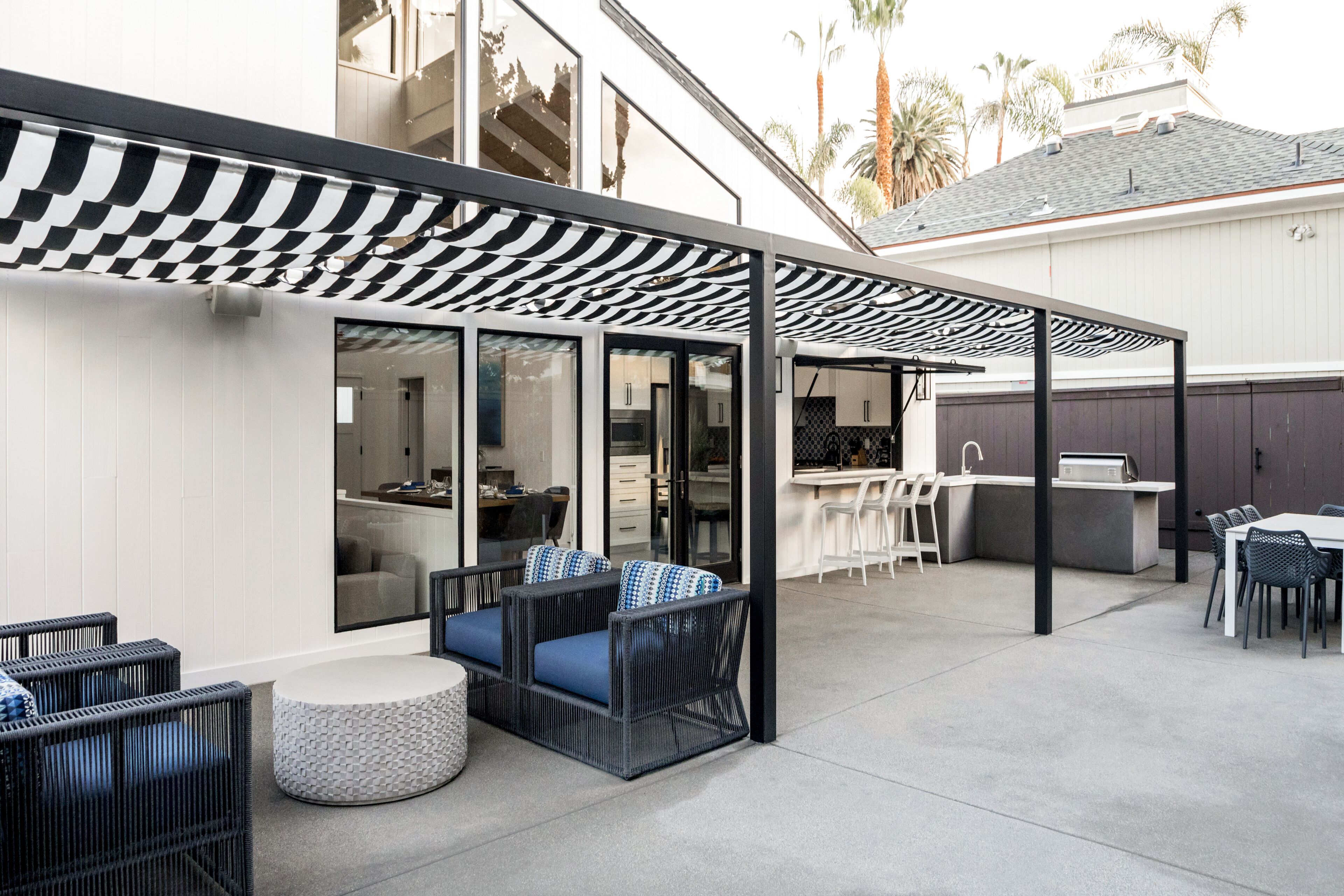 Un patio al aire libre con toldo a cuadros, mobiliario exterior con cojines y una cocina visible a través de puertas de vidrio.