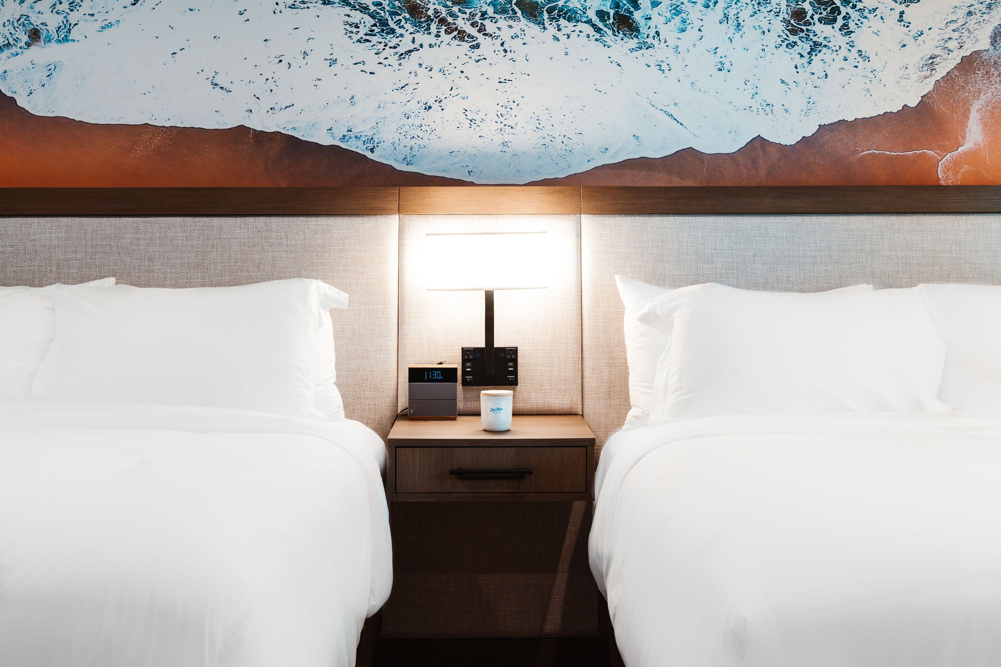 Een hotelkamer met twee bedden, een nachtkastje met een klok en een koffiezetapparaat, en een muurschildering.
