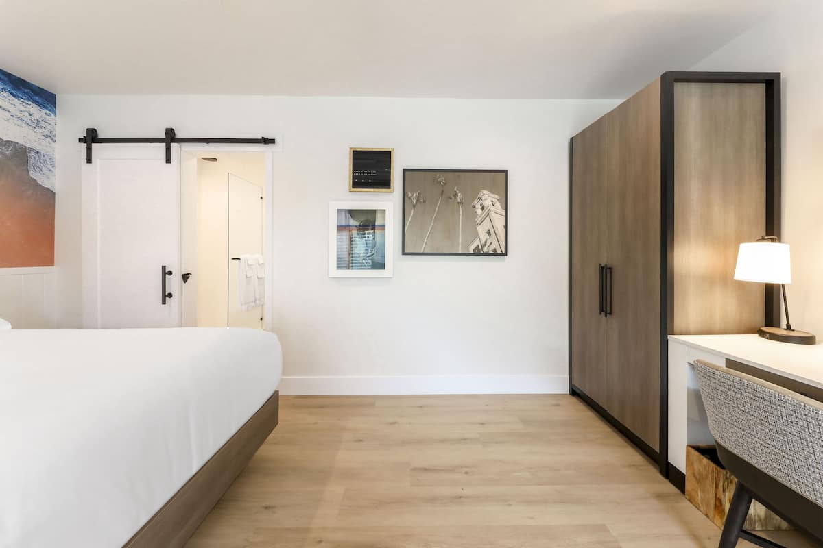 A hotel room with a bed, a desk, and a wardrobe. There are wall-mounted artworks and a door leading to another room.