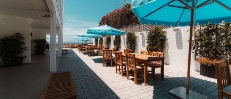 An outdoor patio with wooden tables and chairs, blue umbrellas, and potted plants.