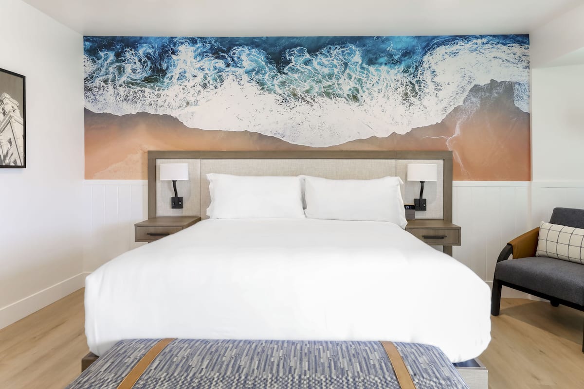 A hotel room with a large ocean wave mural on the wall, a bed with white linens, two bedside tables with lamps, and a chair with a cushion.
