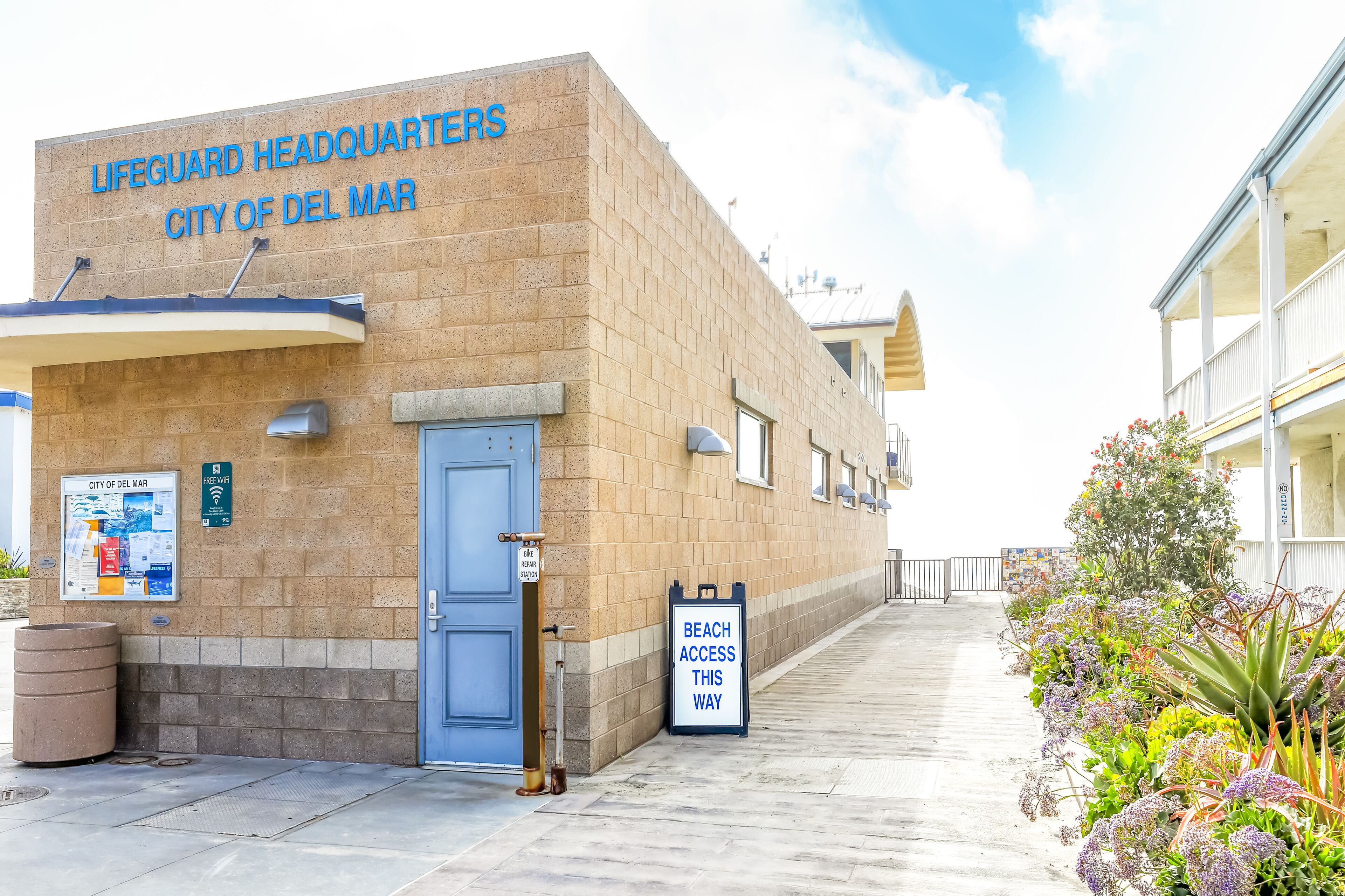 A building labeled "Lifeguard Headquarters City of Del Mar" with a blue door, a trash can, and a sign indicating beach access.