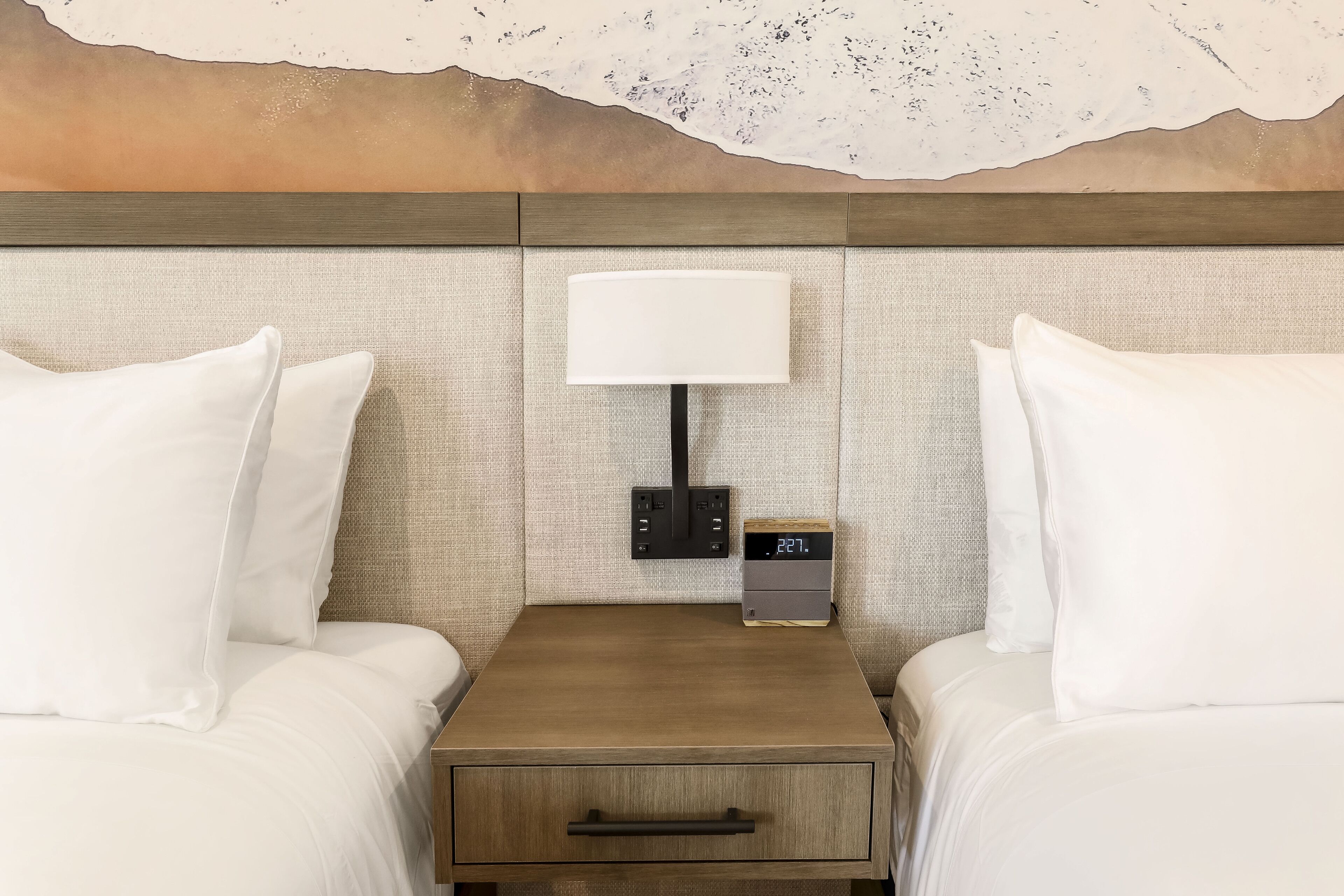 A hotel room with two beds, a nightstand with a lamp, a digital clock, and a wall-mounted artwork.
