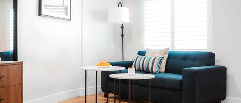 Ein modernes Wohnzimmer mit einem dunkelblauen Sofa, zwei kleinen weißen Tischen, einer Stehlampe und einer Holzkommode.