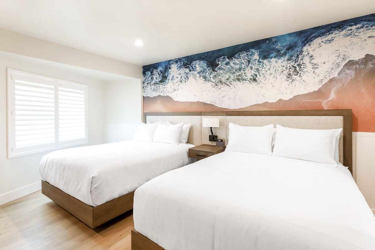A hotel room with two beds, a large wave painting on the wall, a window with blinds, and a bedside table with a lamp.