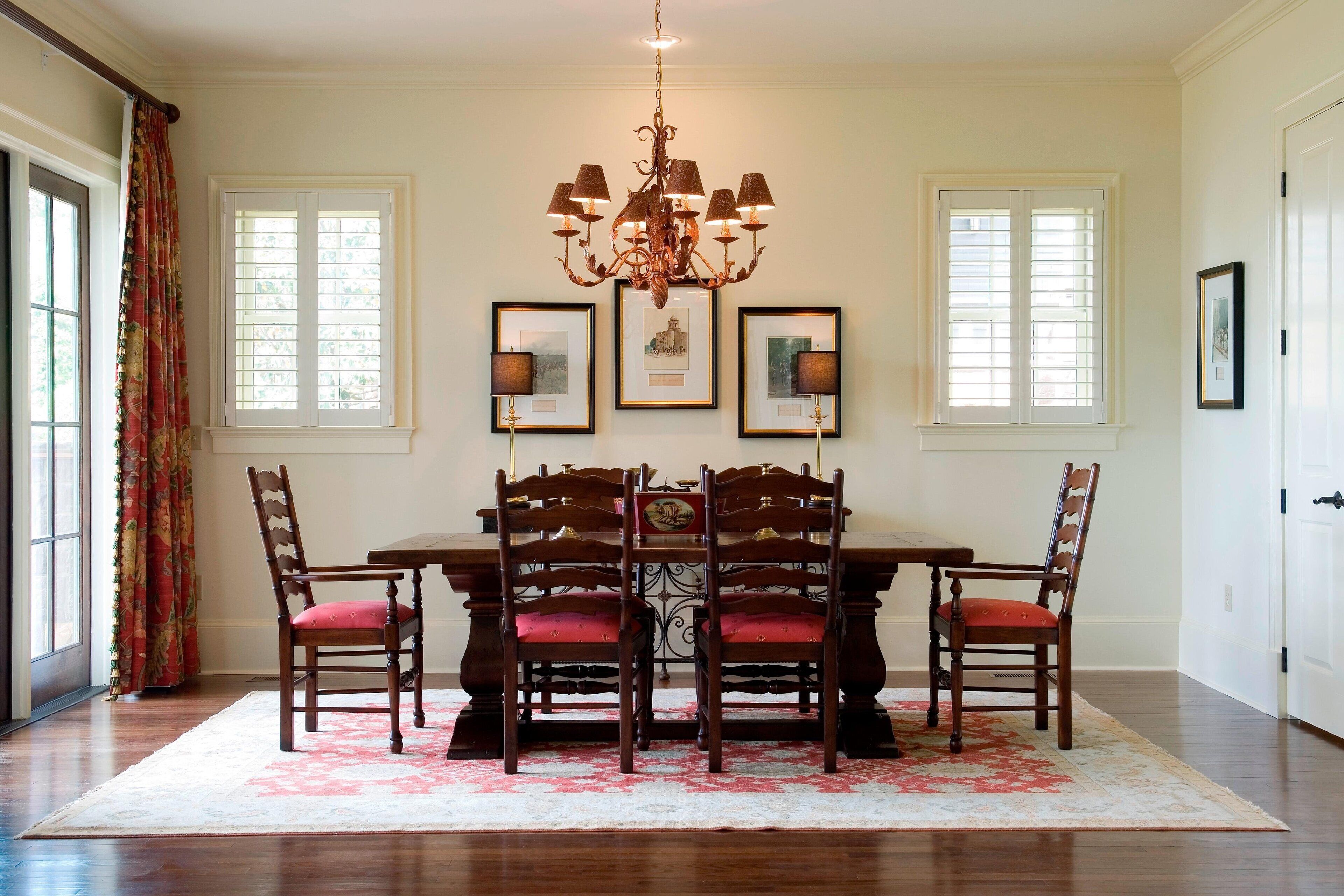 A dining room with a wooden table and chairs, a chandelier, framed pictures, and large windows with curtains.