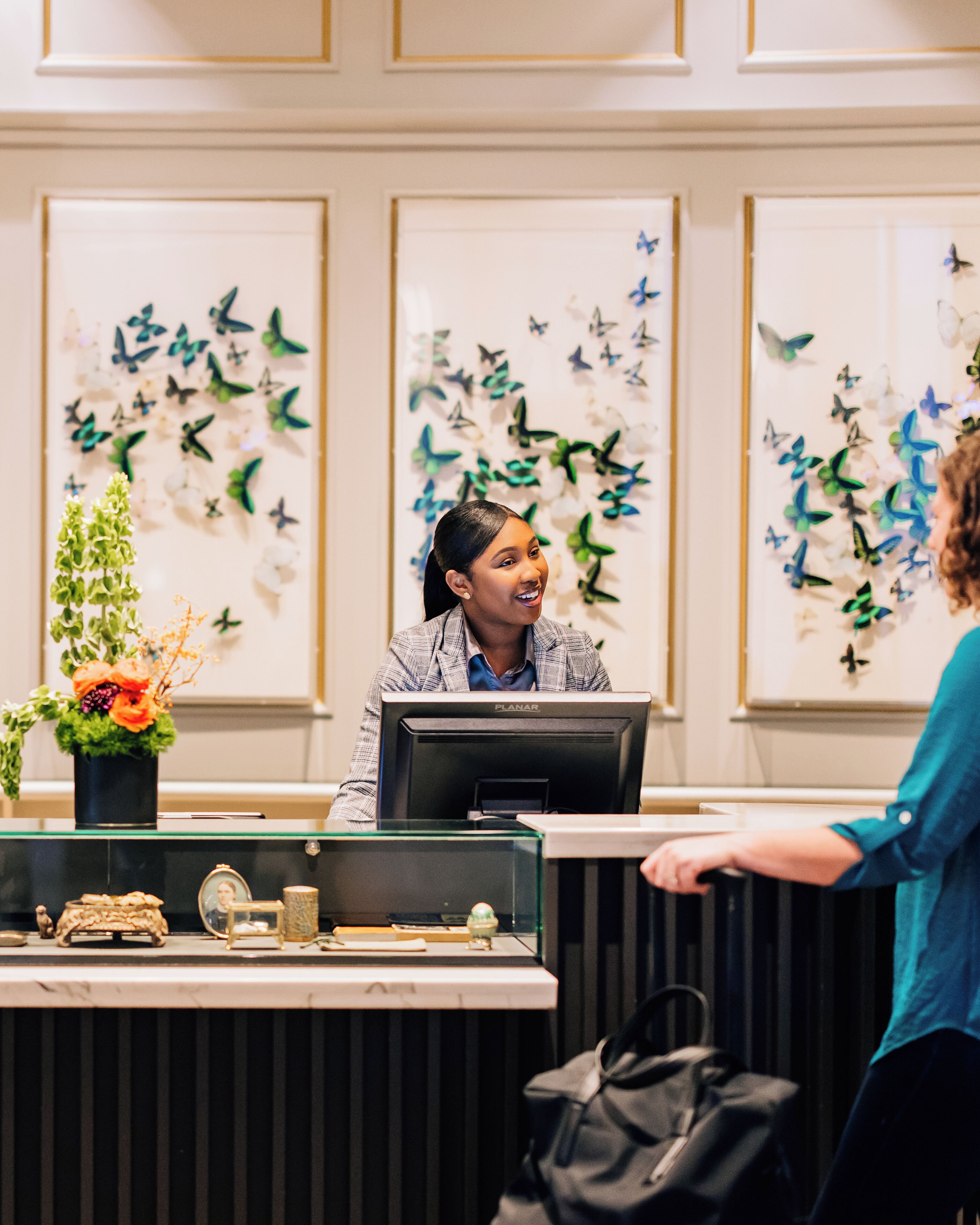 A reception desk with a computer, a small plant, and decorative items. There are butterfly wall decorations and a framed picture on the wall behind the desk.
