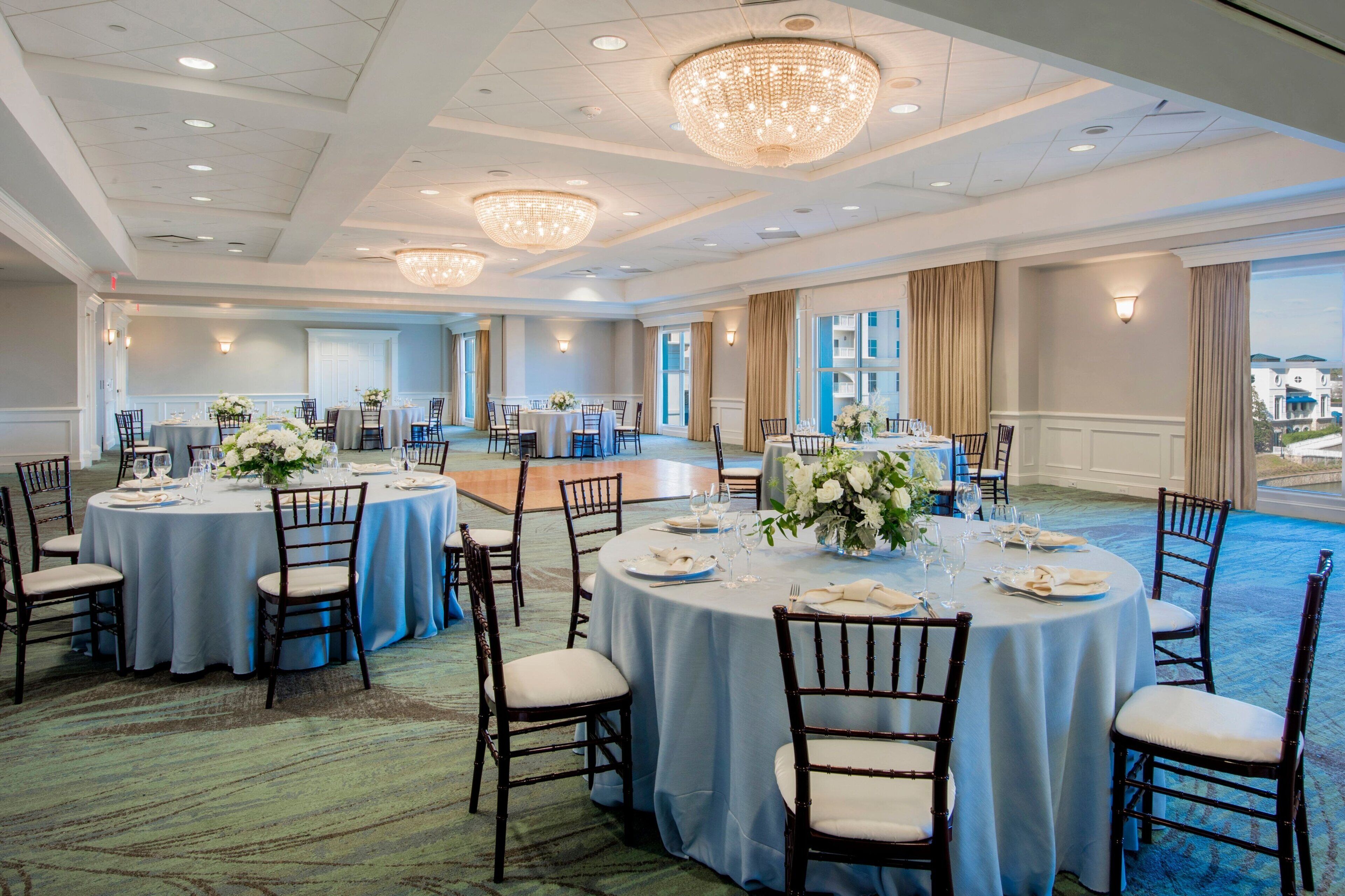 A spacious banquet hall with round tables, elegant chandeliers, and a carpeted floor.
