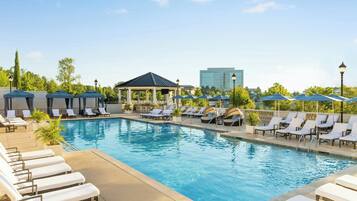 A pool area with lounge chairs and umbrellas, a gazebo, and a modern building in the background.