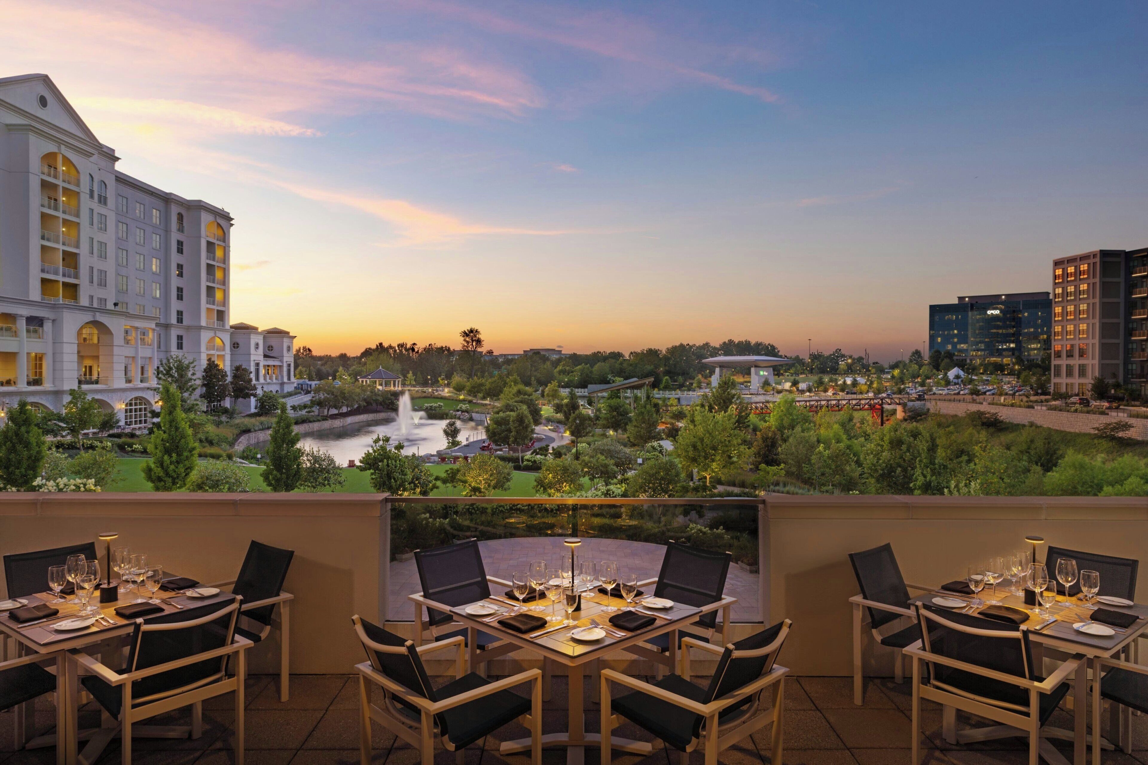 Ein Restaurant mit Außenbereich, das auf einen angelegten Garten und einen Fluss mit Gebäuden im Hintergrund blickt.