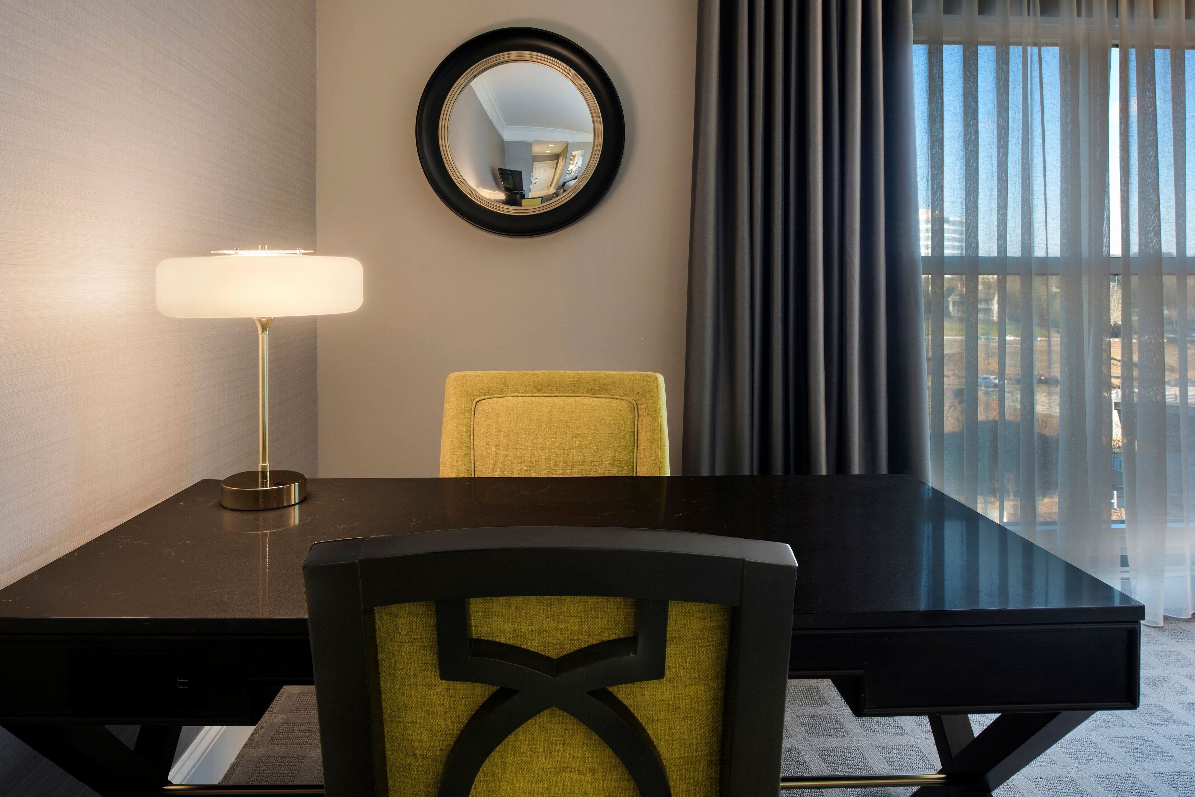 Ein Hotelzimmer mit Schreibtisch, Stuhl, Lampe, Spiegel und Blick ins Freie durch ein Fenster.