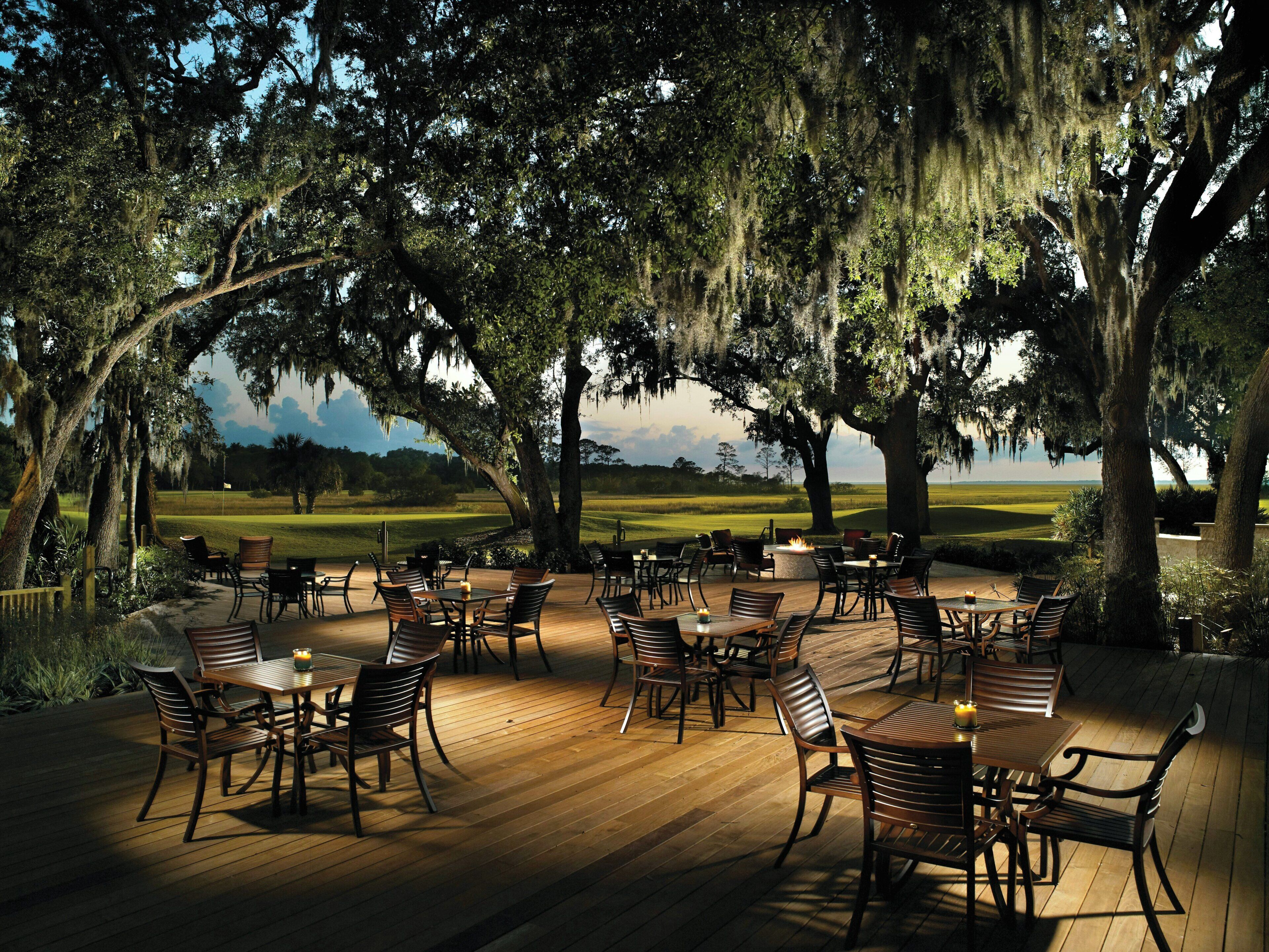 Un área de comedor al aire libre con mesas y sillas de madera, rodeada de árboles grandes con musgo colgante, con vistas a un campo de golf.