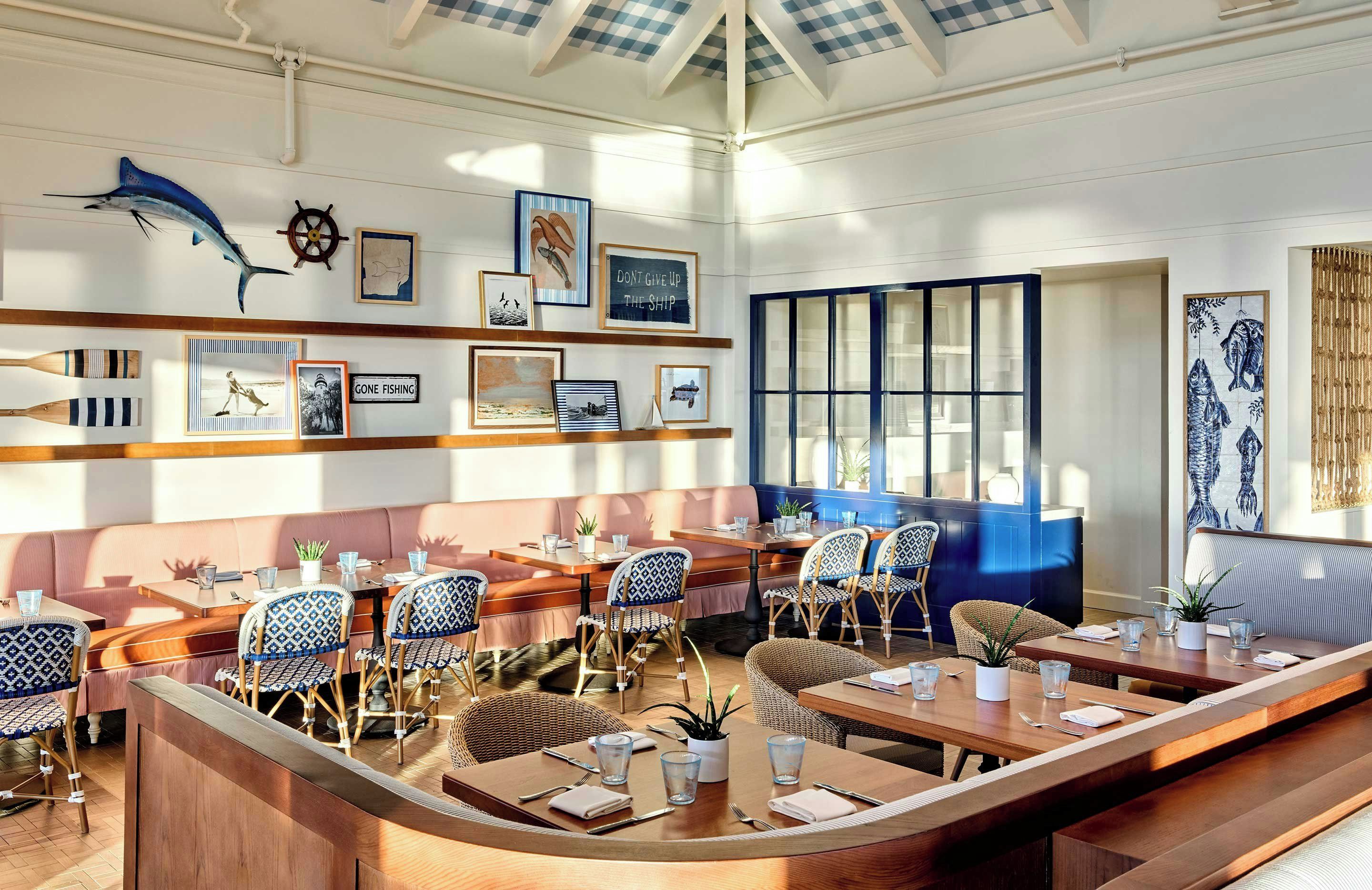 Un restaurant avec des tables et des chaises en bois, une grande fenêtre et des œuvres d’art murales représentant un poisson et un gouvernail de navire.