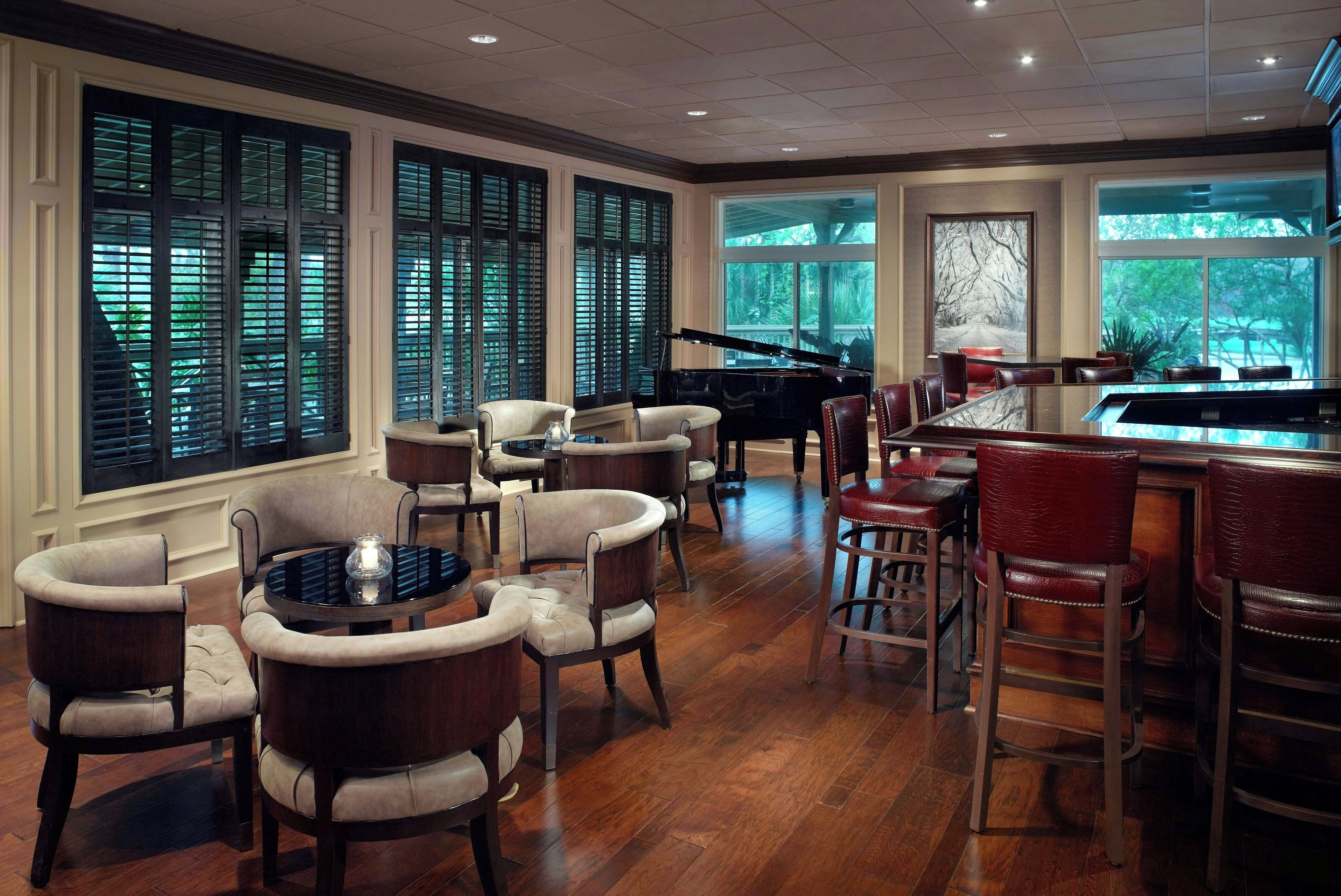 A spacious room with a grand piano, leather chairs, a round table, and large windows with blinds.