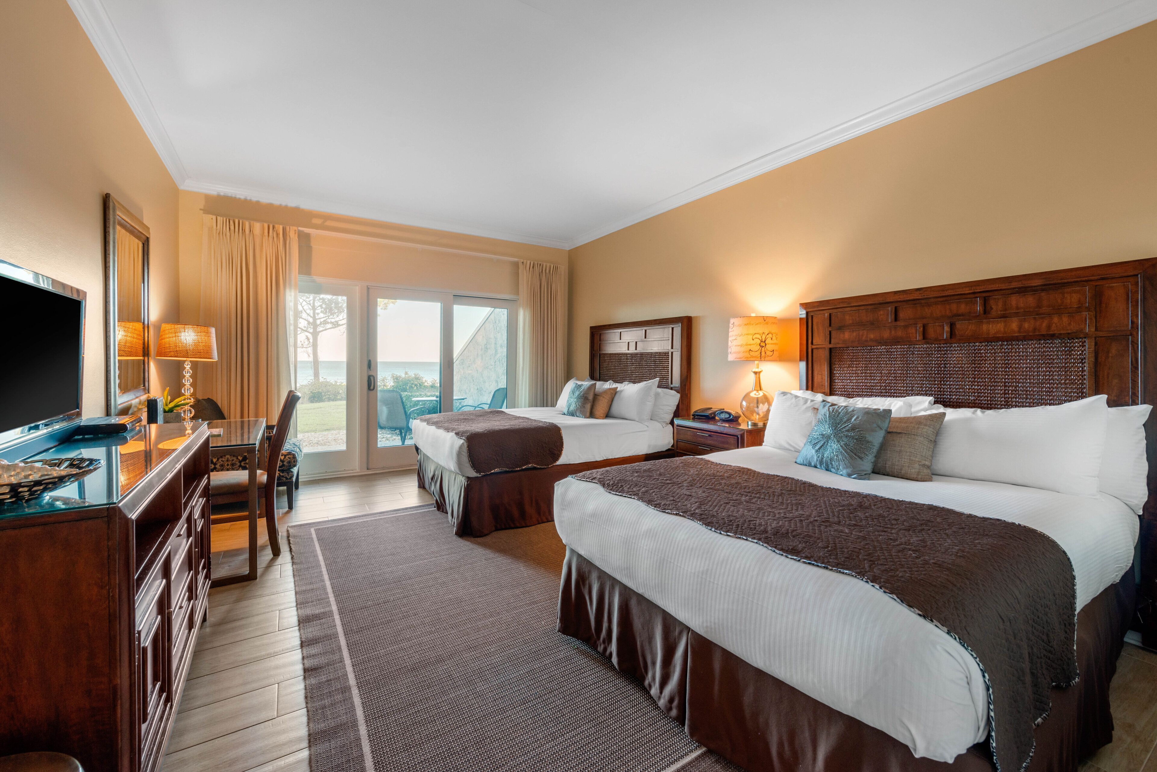 A hotel room with two beds, a desk, a TV, and a balcony with a view.