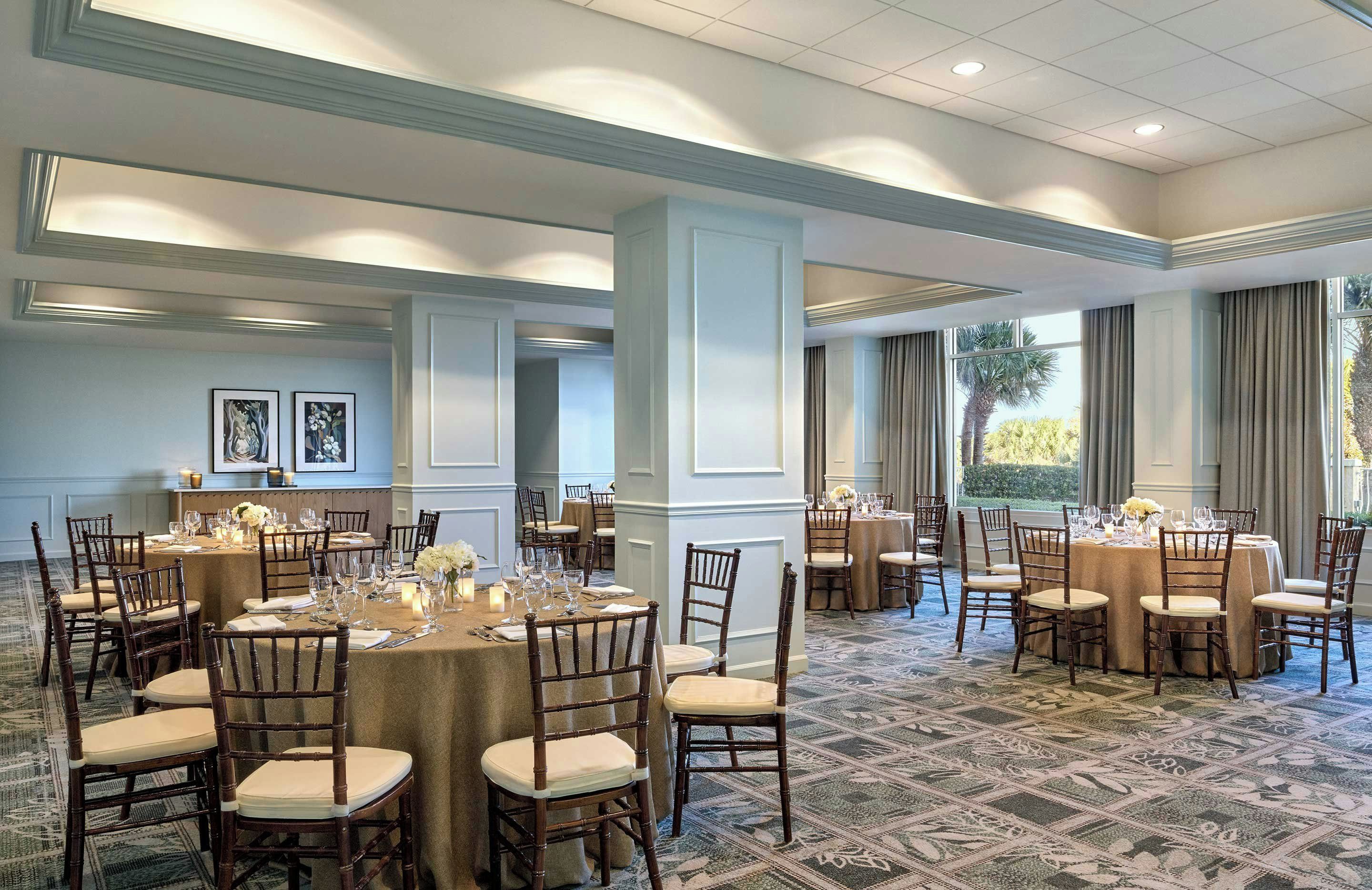 A hotel dining area with multiple round tables set for guests, featuring wooden chairs and beige tablecloths.