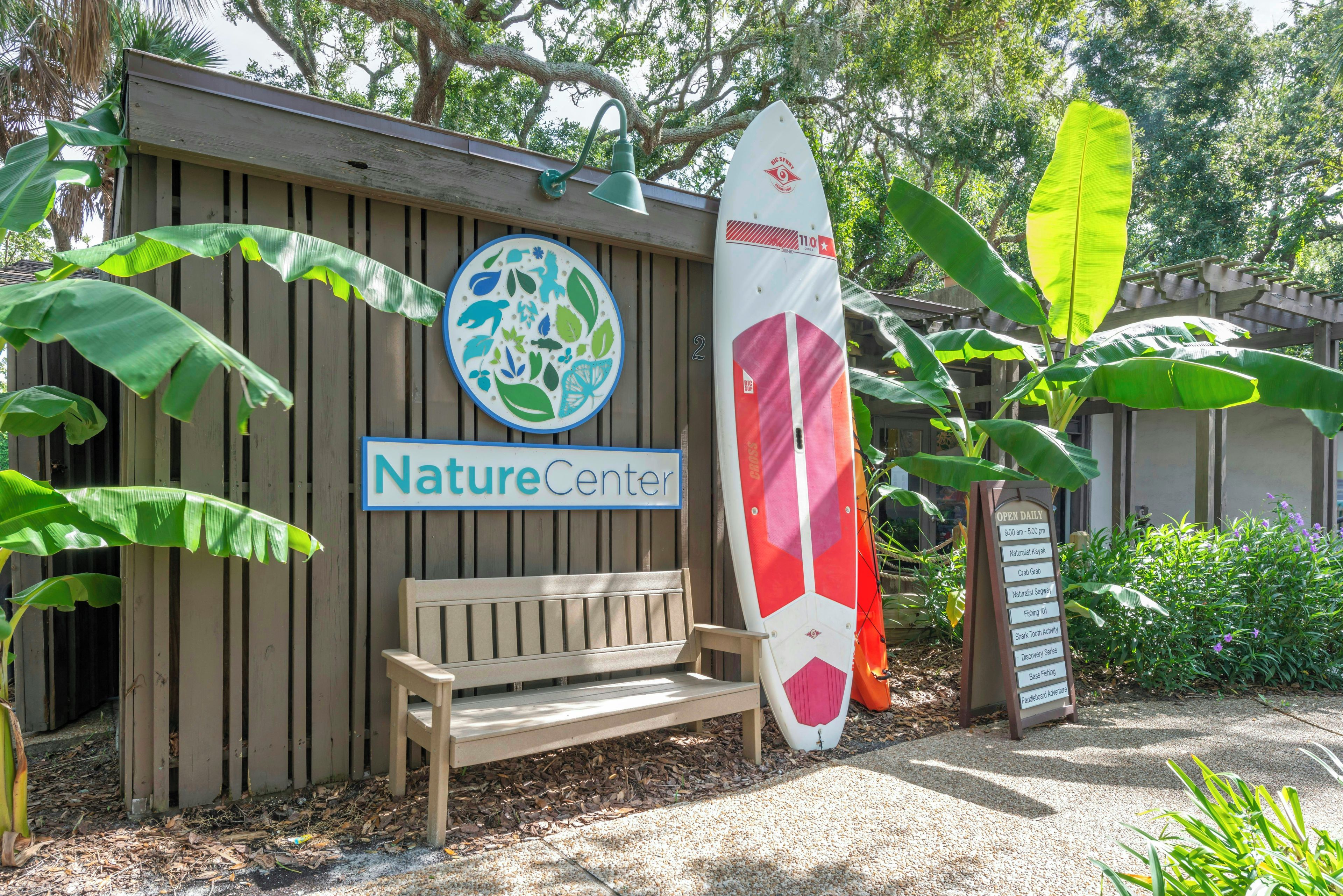 A wooden bench, a surfboard, a Nature Center sign, and a building with a wooden facade.