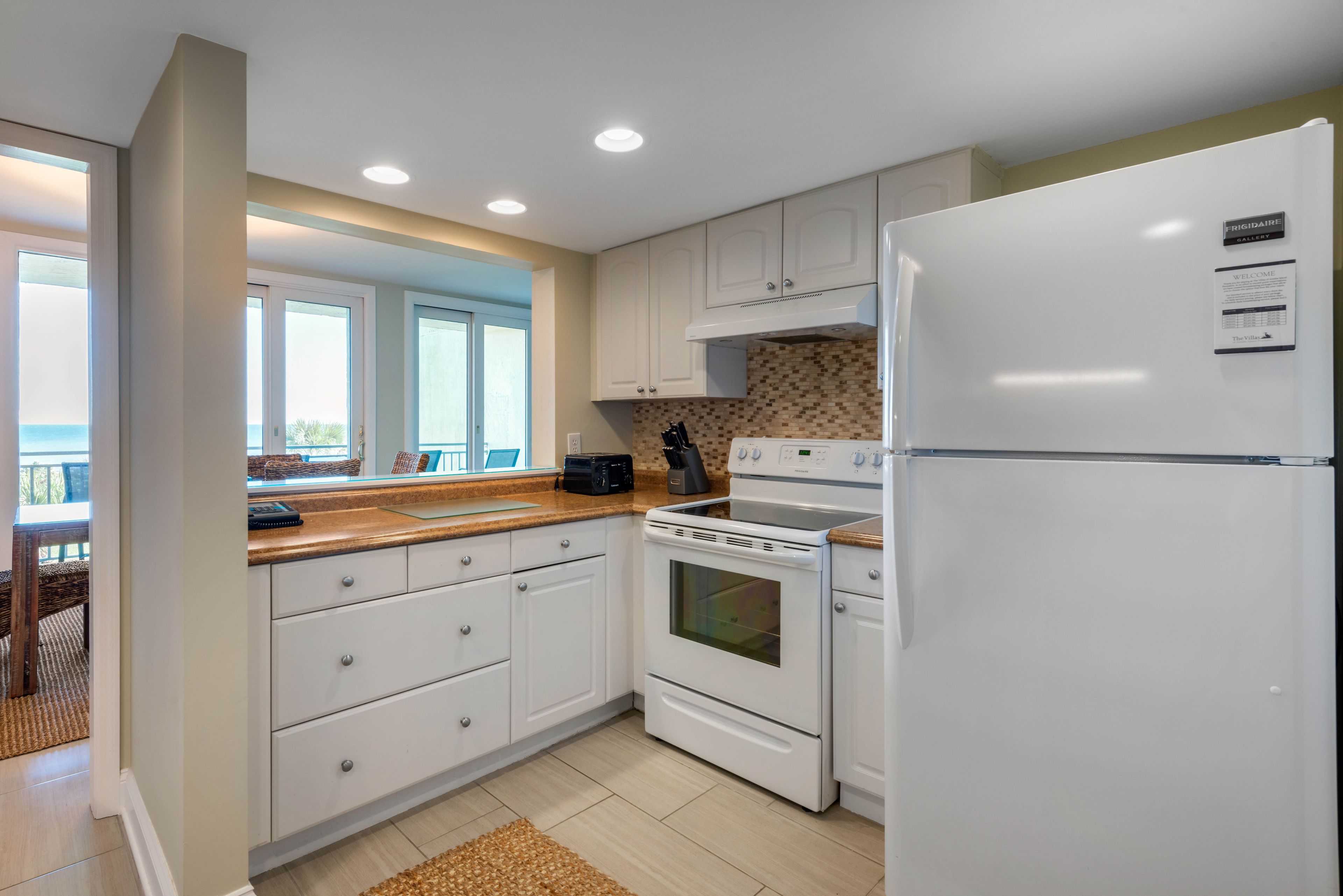 Una cocina moderna con armarios blancos, horno empotrado, refrigerador y ventana con vista a un balcón.
