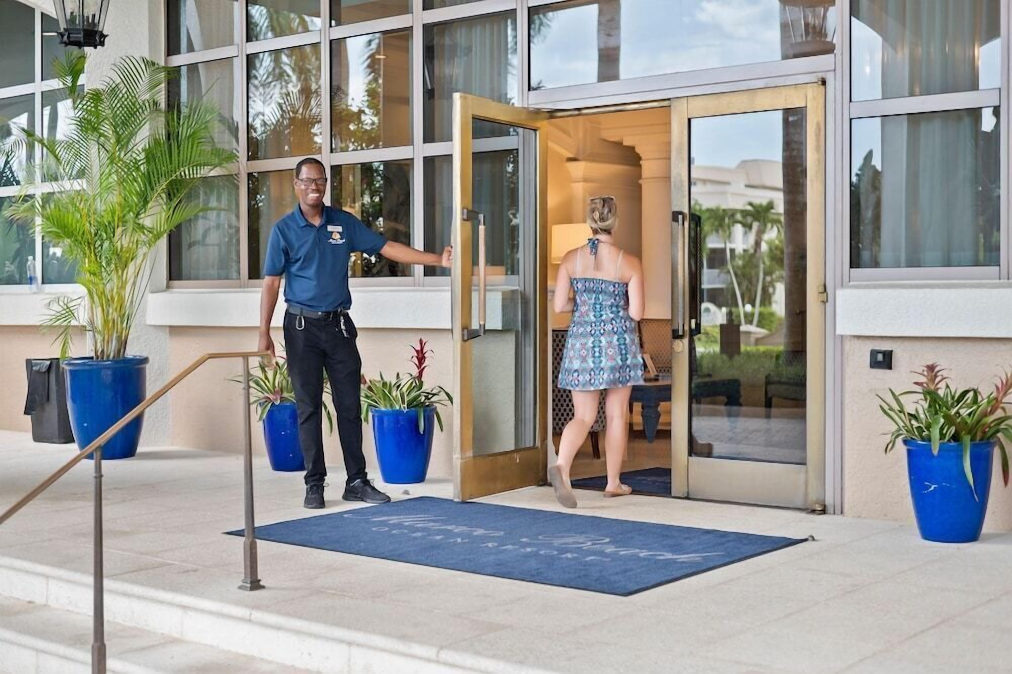 Ein Hotel Eingang mit einem Mann in Uniform an der Tür, einer Frau in einem Kleid, die hereinkommt, und Blumentöpfen ringsum.