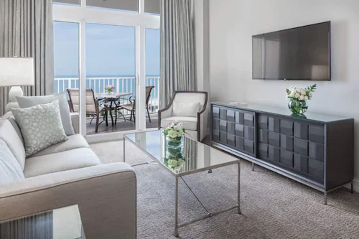 Ett modernt vardagsrum med en soffa, ett soffbord i glas, en tv som är monterad på väggen och en balkong med utsikt över havet.
