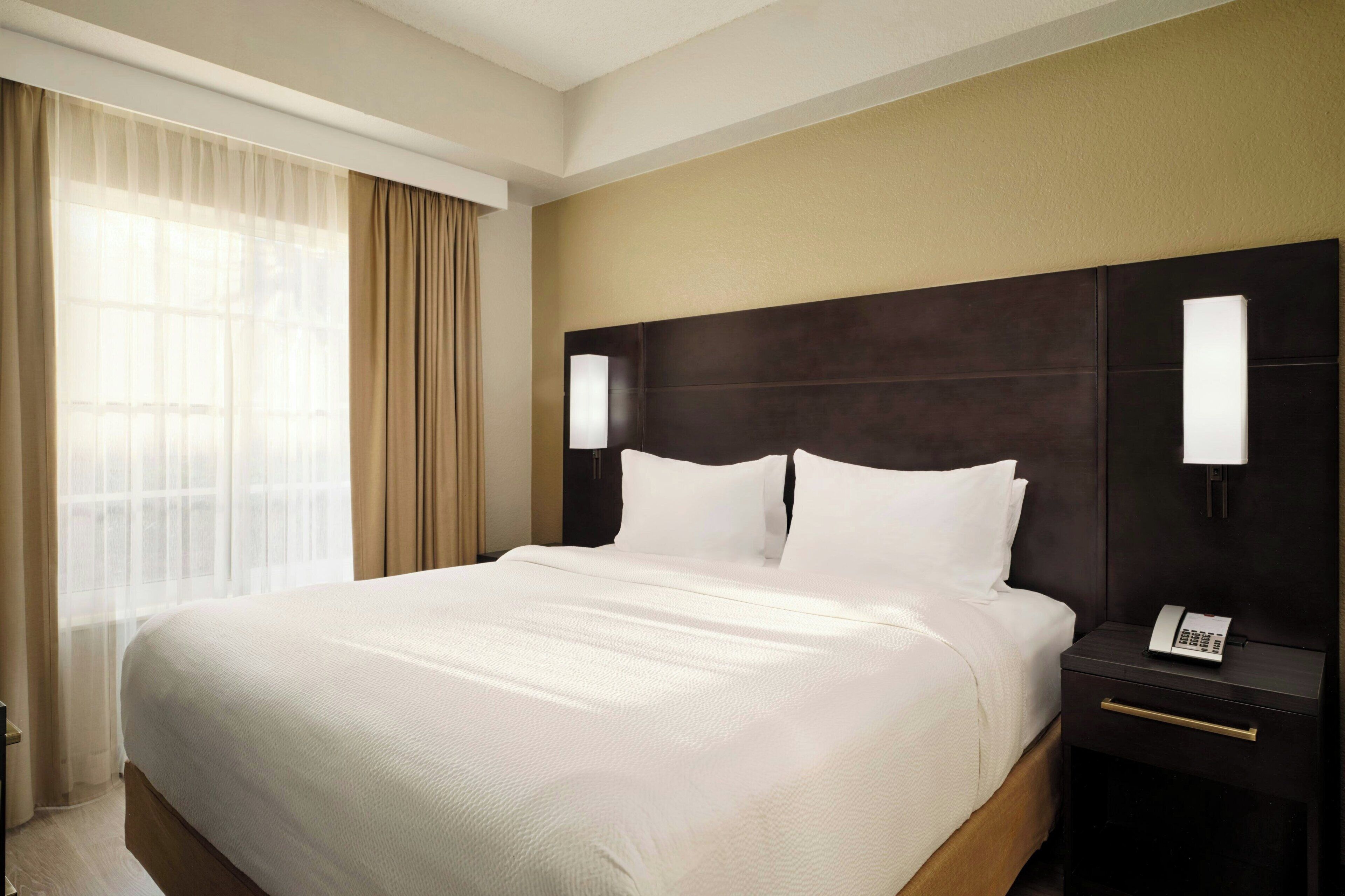 Ein Hotelzimmer mit einem großen Bett, einem Nachttisch mit Telefon und einem Fenster mit Vorhängen.