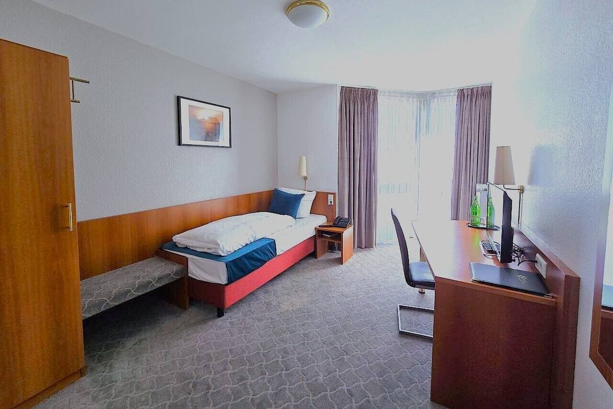A hotel room with a bed, a desk, a chair, a lamp, a picture on the wall, and a window with curtains.